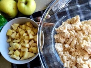 A mixing bowl with dough, a small bowl with sliced ​​apples, an apple next to the bowl and a cinnamon stick.