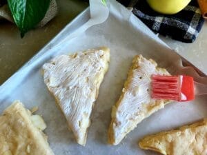 Few scones on a baking tray being brushed with whipping cream. 