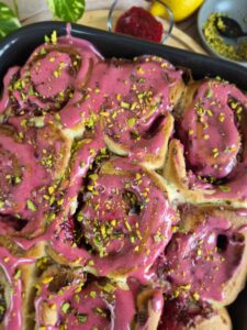 Close-up of golden baked Raspberry Rose Rolls topped with raspberry glaze and sprinkled with chopped pistachios.