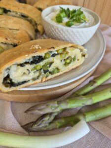 Slice of golden baked Spring Veggie Roll next to fresh asparagus and a bowl of a dip.